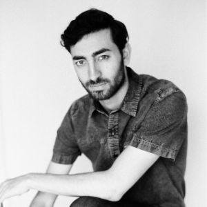 Brandon Powers: Black-and-white portrait of Brandon Powers, a man with dark wavy hair and a trimmed beard, wearing a short-sleeved button-up shirt, looking directly at the camera with a neutral expression.
