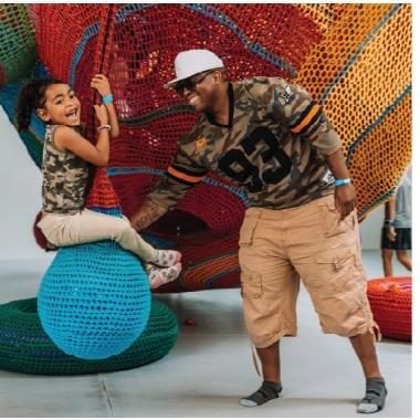 A father dressed in military clothes with child swinging on a crocheted ball in an art installation.