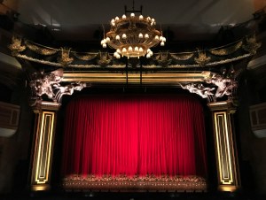 An ornate proscenium theater with a red curtain seen from the audience.
